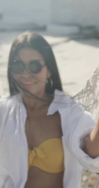 Güneş gözlüklü mutlu, çift ırklı kadının hamakta ağır çekimde yatışının dikey videosu. Yaz, rahatlama, iletişim ve tatiller.