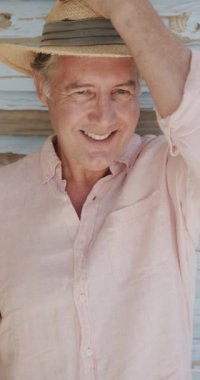 Ahşap duvara karşı şapka takan yaşlı beyaz adamın dikey video portresi, yavaş çekim. Emeklilik, rahatlama, tatiller ve yaşlı yaşam tarzı.