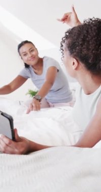 Çift ırklı mutlu çiftin yatakta yavaş çekimde konuştuğu ve tablet kullandığı dikey video. Ev hayatı, yaşam tarzı ve aşk..