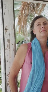 Sahil evinin verandasında dinlenen mutlu beyaz bir kadının dikey video portresi, yavaş çekim. Emeklilik, rahatlama, tatiller ve yaşlı yaşam tarzı.