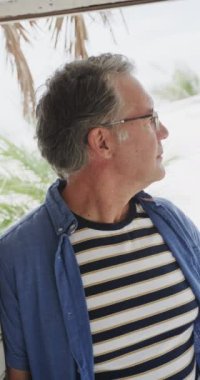 Sahil evinin verandasından seyreden mutlu, yaşlı beyaz bir adamın dikey videosu, yavaş çekim. Emeklilik, rahatlama, tatiller ve yaşlı yaşam tarzı.