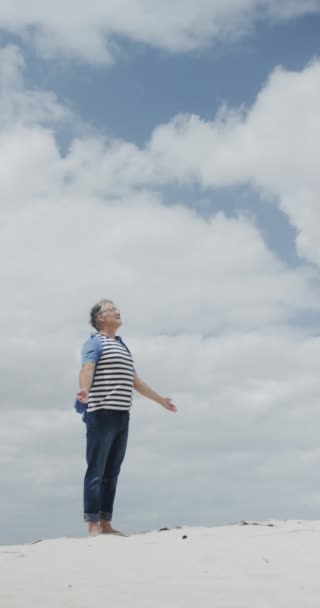 一个快乐的老年高加索人赤脚在海滩上走路 举起胳膊 慢动作的垂直录像 休假和老年生活方式 — 图库视频影像