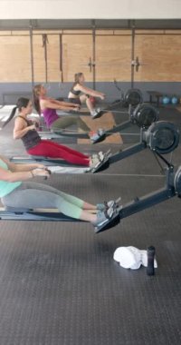 Spor salonundaki kürek makineleriyle yavaş çekimde egzersiz yapan çeşitli sporcu kadınların dikey videosu. Spor, spor, sağlıklı yaşam tarzı ve etkinlik.