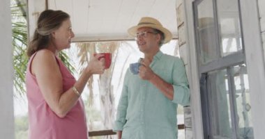 Kahvelerini yudumlayan ve verandadaki sahil evinde ağır çekimde konuşan mutlu beyaz çift. Emeklilik, rahatlama, tatiller ve yaşlı yaşam tarzı.