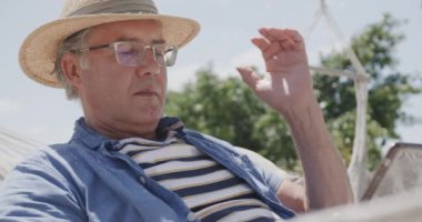 Güneş şapkalı, kumsalda tablet kullanan, ağır çekimde hamakta oturan mutlu beyaz adam. Emeklilik, rahatlama, iletişim, tatiller ve kıdemli yaşam tarzı.