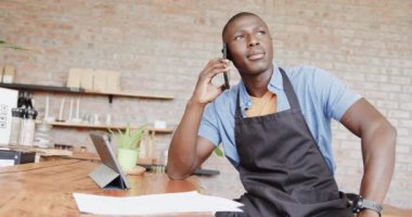 Afroamerikan erkek barista tablet kullanıyor ve kafede akıllı telefondan konuşuyor, ağır çekimde. Yaşam tarzı, iletişim, girişimcilik ve yerel işletme.