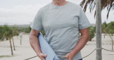 Elinde yoga minderi tutan ve kumsalda ağır çekimde gülümseyen yaşlı beyaz adamın portresi. Emeklilik, dinlenme, tatiller ve sağlıklı yaşlı yaşam tarzı..