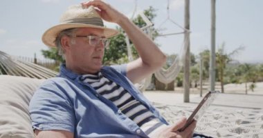 Hamakta ağır çekimde akıllı telefon kullanan, güneş şapkalı, ciddi bir beyaz adam. Emeklilik, rahatlama, iletişim, tatiller ve kıdemli yaşam tarzı.