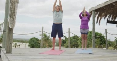 Kumsalda, güneş güvertesinde ağır çekimde yoga yapan son sınıf beyaz çift. Emeklilik, dinlenme, tatiller ve sağlıklı yaşlı yaşam tarzı..