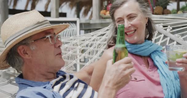 幸せな高齢者の白人のカップルを飲むとビーチバーの外のハンモックで ゆっくりと動きで話しています リラクゼーション ロマンス シニアライフスタイル — ストック動画
