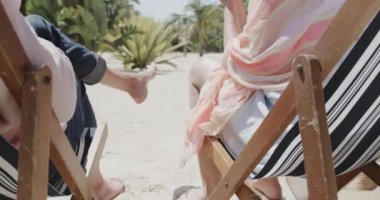 Kumsaldaki güverte sandalyelerinde oturan ve ağır çekimde bira içmeye çalışan mutlu beyaz çift. Emeklilik, rahatlama, birliktelik, tatiller ve kıdemli yaşam tarzı.