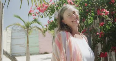 Yavaş çekimde pembe çiçeklerle güneşe gülümseyen son sınıf beyaz kadın portresi. Emeklilik, rahatlama, tatiller ve yaşlı yaşam tarzı.