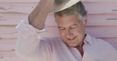 Yaşlı beyaz bir adamın portresi ağır çekimde yıpranmış pembe ahşap duvarla şapka takıyor. Emeklilik, rahatlama, tatiller ve yaşlı yaşam tarzı.