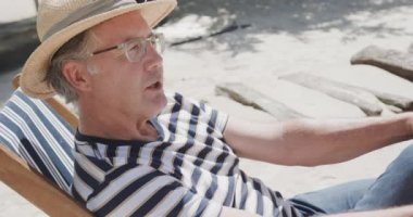 Güverte sandalyesinde oturan, güneşli sahilde ağır çekimde konuşan kıdemli beyaz adam. Emeklilik, rahatlama, tatiller ve yaşlı yaşam tarzı.