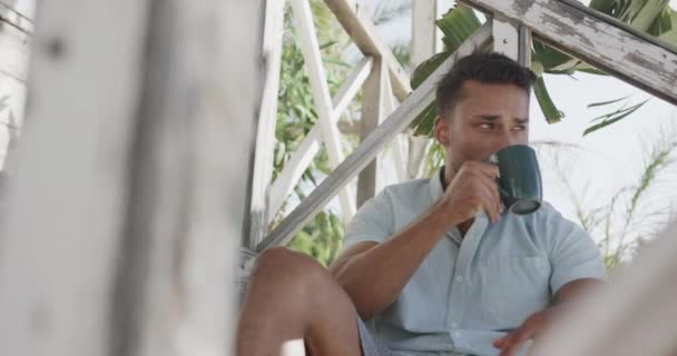 Szczęśliwy Dwurasowy Mężczyzna Pijący Kawę Ganku Domku Plaży Zwolnionym Tempie — Wideo stockowe