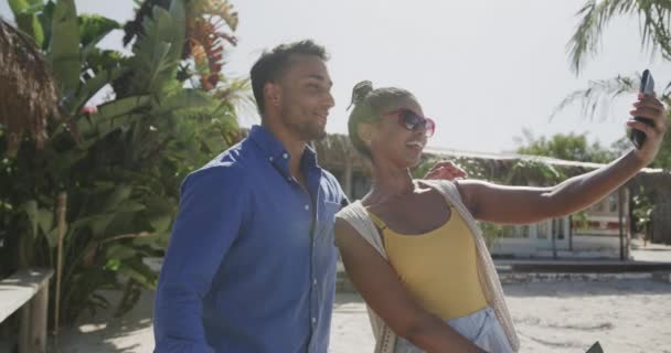 这对快乐的夫妻面带微笑 带着智能手机在阳光明媚的海滩上慢吞吞地走着 交流和生活方式 — 图库视频影像