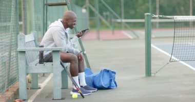 Mutlu Afro-Amerikan adam akıllı telefonuyla konuşuyor tenis kortunda bankta oturuyor, ağır çekimde. Spor, hafta sonu, iletişim, eğlence ve etkinlik.