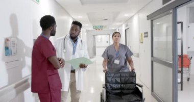 Odaklanmış farklı erkek doktorlar konuşuyor ve tekerlekli sandalyeli kadın hemşire ağır çekimde, değişmemiş. Sağlık, ilaç ve hastane koridoru..