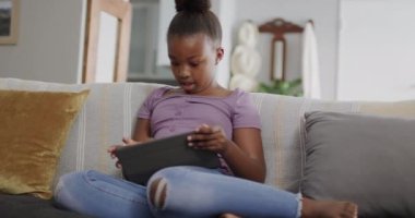 Afro-Amerikalı kız kanepede oturuyor ve tablet kullanıyor, ağır çekimde. Yaşam tarzı, iletişim, çocukluk ve ev hayatı.