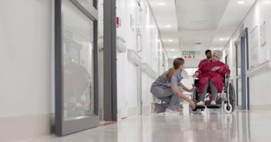 Ağır çekimde, değişmeden tekerlekli sandalyeli yaşlı bir hastayla yürüyen çok çeşitli kadın hemşireler. Sağlık, ilaç ve hastane koridoru..