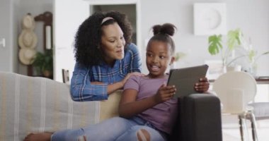 Mutlu Afro-Amerikan anne ve kızı kanepede oturuyor, tablet kullanıyor, ağır çekimde. Yaşam tarzı, iletişim, aile ve aile hayatı.