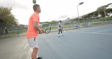 İki farklı erkek arkadaş, açık hava tenis kortunda ağır çekimde çift top oynuyorlar. Spor, arkadaşlık, takım çalışması, eğlence, hafta sonu, yaşam tarzı ve etkinlik.