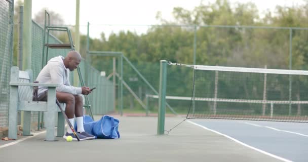 Hombre Afroamericano Feliz Usando Teléfono Inteligente Sentado Banco Cancha Tenis — Vídeo de stock