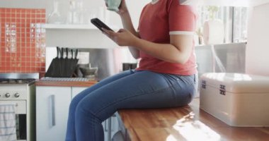 Kahve içen ve evde ağır çekimde akıllı telefon kullanan mutlu çift ırklı kadın. Ev hayatı, yaşam tarzı ve teknoloji.