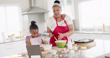 Değiştirilmemiş Afro-Amerikan anne ve kızı, mutfakta tablet kullanarak yavaş çekimde yemek pişiriyorlar. Yemek pişirme, yaşam tarzı, iletişim, aile, birliktelik ve ev hayatı.