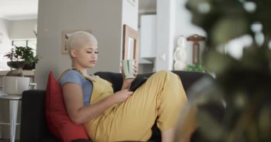 Akıllı telefon kullanıp kanepede ağır çekimde kahveyle oturan mutlu melez bir kadın. Ev hayatı, yaşam tarzı ve teknoloji.