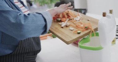 Mutfakta sebze atıkları taşıyan önlüklü melez bir kadının orta kesimi, ağır çekim. Yemek, yemek, sağlıklı yaşam, ekoloji, geri dönüşüm ve ev hayatı.
