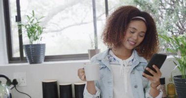 Akıllı telefon kullanan ve mutfakta kahve içen mutlu, melez bir kadın. İletişim, yaşam tarzı, rahatlama ve ev hayatı, değişmemiş..