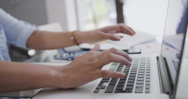 Evde ağır çekimde dizüstü bilgisayar ve akıllı telefon kullanan melez bir kadının orta kısmı. Uzaktan çalışma, iletişim, yaşam tarzı ve evden çalışma.