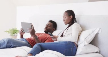 Afro-Amerikalı mutlu çift yatakta uzanıp konuşuyorlar, akıllı telefon ve tablet kullanıyorlar, ağır çekimde. Yaşam tarzı, ilişki, rahatlama, iletişim, birliktelik ve aile hayatı, değişmemiş..