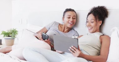 Çift ırklı mutlu lezbiyen çift tablet ve akıllı telefon kullanarak yatakta oturuyor ve yavaş çekimde konuşuyor. Yaşam tarzı, ilişki, birliktelik, rahatlama, iletişim ve ev hayatı, değişmemiş..
