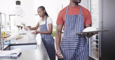 Önlüklü, ekmek pişiren, hamuru fırına koyan mutlu Afro-Amerikan çifti, yavaş çekimde. Yaşam tarzı, yemek pişirme, ekmek, birliktelik, yemek ve ev hayatı, değişmemiş.