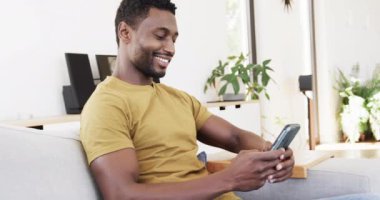 Mutlu Afro-Amerikan adam akıllı telefon kullanıyor oturma odasında koltukta oturuyor, ağır çekimde. Yaşam tarzı, rahatlama, iletişim ve ev hayatı, değişmemiş..