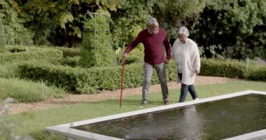 Üst düzey melez çift el ele tutuşup güneşli bahçede birlikte yürüyorlar, değişmeden, ağır çekimde. Kıdemli yaşam tarzı, yaz, doğa, boş zaman ve ev hayatı..