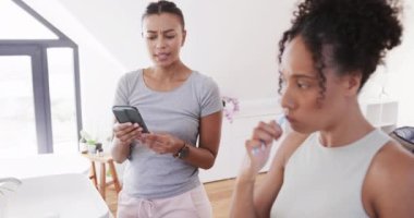 Çift ırklı mutlu lezbiyen çift dişlerini fırçalıyor ve güneşli banyoda akıllı telefon kullanıyorlar, ağır çekimde. Yaşam tarzı, ilişki, birliktelik, kendine bakım, iletişim ve ev hayatı, değişmemiş..