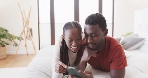 幸せなアフリカ系アメリカ人のカップルベッドの上でスマートフォンを使用して横になり ゆっくりとした動きで自宅で話しています リラクゼーション コミュニケーション 一緒に 愛と国内生活 変わらない — ストック動画