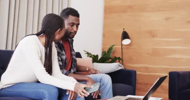 幸せなアフリカ系アメリカ人のカップルは スマートフォンやノートパソコンを使ってゆっくりとソファで話しています ライフスタイル コミュニケーション テクノロジー 国内生活 — ストック動画