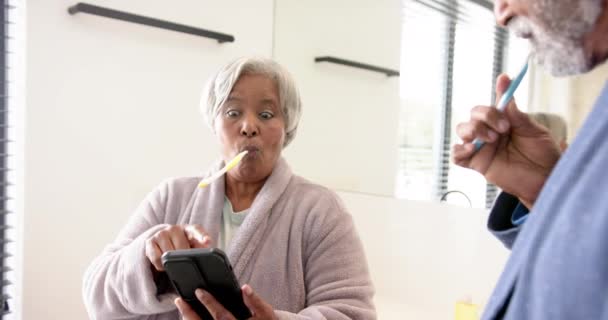 Feliz Casal Biracial Sênior Escovando Dentes Usando Smartphone Banheiro Inalterado — Vídeo de Stock