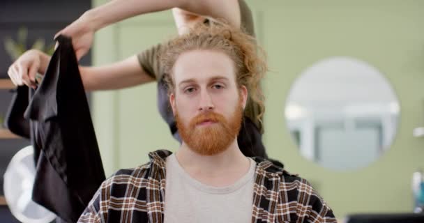 幸せな白人女性理容師は ゆっくりと動きながら 理髪店でひげを生やした男性のクライアントにケープを置きます 地元のビジネス サービス ヘアケアとヘアスタイル 変更なし — ストック動画
