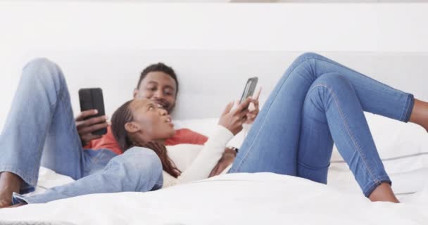 幸せなアフリカ系アメリカ人のカップルのベッドで話をし スマートフォンを使用して横たわっ スローモーション リラクゼーション コミュニケーション 一緒に 愛と国内生活 変わらない — ストック動画