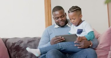 Afro-Amerikan baba ve oğlu kanepede ağır çekimde tablet ve beşlik çakarak oturuyorlar. Aile, iletişim, birliktelik ve aile hayatı değişmedi..