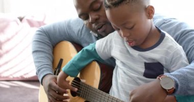 Mutlu Afro-Amerikan baba ve oğlu kanepede oturup gitar çalıyor, ağır çekimde. Müzik, aile, yaratıcılık, öğrenme, keşif ve birliktelik, değiştirilmemiş.