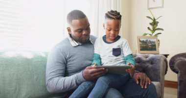 Mutlu Afro-Amerikan babası koltukta oğlunun kucağında ağır çekimde tablet kullanarak oturuyor. Aile, iletişim, birliktelik ve aile hayatı değişmedi..