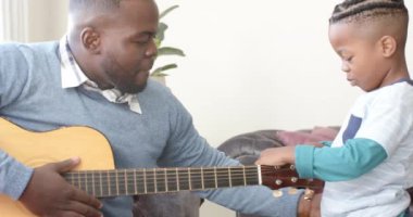 Mutlu Afro-Amerikan baba ve oğlu kanepede oturup gitar çalıyor, ağır çekimde. Müzik, aile, yaratıcılık, öğrenme, keşif ve birliktelik, değiştirilmemiş.