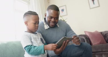 Mutlu Afro-Amerikan baba ve oğlu kanepede oturup ağır çekimde, oturma odasında tablet kullanıyor. Aile, iletişim, birliktelik ve aile hayatı değişmedi..