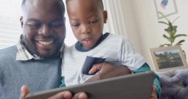 Mutlu Afro-Amerikan babası koltukta oğlunun kucağında ağır çekimde tablet kullanarak oturuyor. Aile, iletişim, birliktelik ve aile hayatı değişmedi..